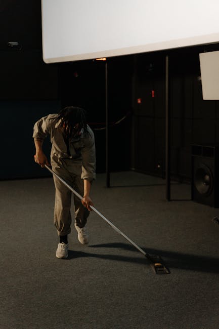 A man wearing a grey jumpsuit and sneakers, operating a vacuum cleaner on a dark carpeted floor in a room with blue walls and tiered seating, performing deep cleaning and sanitisation as part of a com