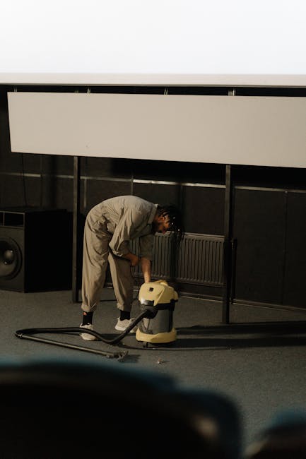 A professional cleaner dressed in full protective gear, including a white coverall, mask, gloves, and shoe covers, is using a steam cleaning tool on a light-colored, textured carpet in a well-lit, mod